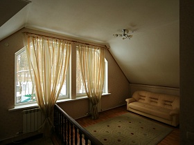 уютный холл второго этажа над лестницей с бежевым диваном, ковром на полу и бежевыми гардинами на окне  съемного двухэтажного дома в сосновом лесу