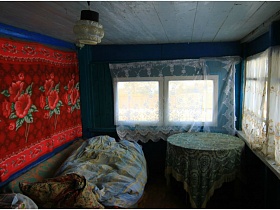 незаправленный диван у синей деревянной стены с плюшевым красным ковром, круглый стол покрытый скатертью у окон веранды с белыми гардинами жилого дома в деревне