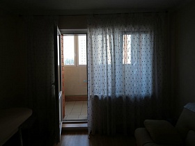 белая с рисунком гардина на окне с открытой дверью на застекленный балкон простой квартиры современной высотки в зеленом массиве, с видом на море