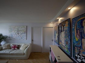 большие картины на стенах над светлым мягким диваном и над бежевым не высоким шкафом в гостиной с белыми стенами и потолком стильной трешки в Икеа стиле