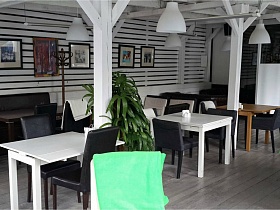 чередование темной и белой мебели в уютном кафе с белыми плафонами светильников на потолке,  деревянные колонны и картины под стеклом на полосатой стене белой веранды