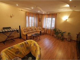 желтый мягкий диван, кресло и журнальный столик в углу гостиной у двух окон с полосатой разноцветной гардиной, детский манеж и комнатные цветы в высоких вазонах на полу простой двухкомнатной квартиры