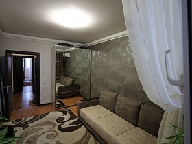 шкаф-купе с зеркальными дверцами, мягкий светлый диван с серыми подушками, светлый с вензелями ковер на полу гостиной из открытого балкона современной трешки