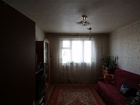 красный цветной ковер на полу между мебельной горкой и мягким диваном с вишневым покрывалом в просторной комнате с большим окном современной трехкомнатной квартиры пенсионеров