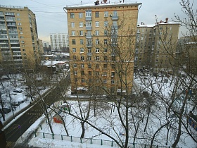 вид из окна двухкомнатной квартиры на соседние многоэтажные дома, придомовую территорию под снегом и автомобильную дорогу