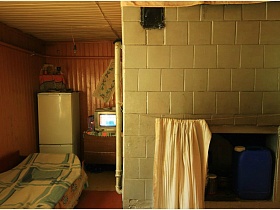 деревянная кровать, застеленная цветным покрывалом, белый холодильник в углу комнаты и печь,облицованная белой плиткой жилого дома в деревне