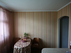 два деревянных стула с подушками на сидушках вокруг обеденного стола с фруктами в розовой плетенной корзине у стены с полосатыми розоватыми обоями на кухне с белой и розовой гардиной на окне