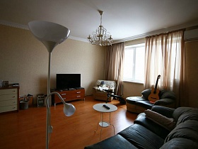 белый торшер, в стиле хай-тек, люстра, стилизованная под свечи на белом потолке гостиной с мягкой мебелью, телевизором, комодом квартиры в переезде(въезде) молодоженов