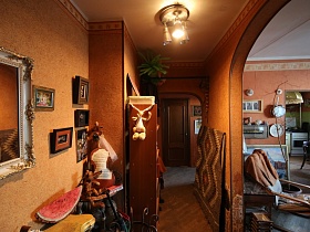 прямоугольное зеркало в белой рамке с инкрустацией, картины в рамках на стене, шкаф и полотно, закрытое ковром в узком коридоре профессорской квартиры в коричневых тонах