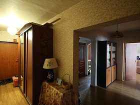 желтый абажур настольной лампы, рамка с фотографией и плетенная корзина на поверхности столика с цветной скатертью, шкаф-купе в длинном коридоре с обувью на коврике у входной двери в современную трехкомнатную квартиру