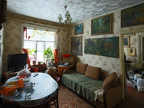 диван с подушками, стулья у круглого  стола с посудой, телевизор на тумбочке у окна и многочисленные картины на стенах столовой деревянной советской дачи художника с овальной террасой