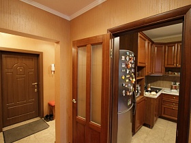 серебристый холодильник, коричневая мебельная кухня через открытую дверь коридора со светло-коричневыми обоями на стенах трехкомнатной квартиры государственного служащего