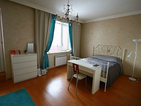 статуэтки на белом комоде, стул у белого деревянного стола возле большой кровати в спальной комнате с серыми и голубыми шторами современной трехкомнатной квартиры