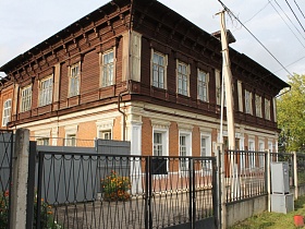 кирпичное здание с достроенным вторым деревянным коричневым этажом старинной школы с серыми воротами за металлическим забором