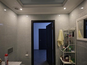 ванные принадлежности и предметы по уходу на полочках белой этажерки в углу ванной комнаты с серой плиткой и натяжным потолком стильной дачки