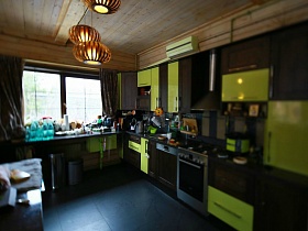 заставленный широкий деревянный подоконник  окна с коричневыми шторами в зоне кухни с коричнево-салатовой мебельной стенкой на сером полу просторной гостиной современного загородного дома