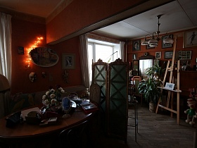 картина,фотографии, маска и лампочки гирлянды на стене у окна кухни с деревянным столом у дивана за ширмой между двумя комнатами трехкомнатной квартиры