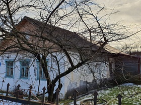 голубые наличники на окнах дома-мазанки с белыми стенами,треугольной крышей, покрытой шифером, пристроенной деревянной верандой на участке за металлическим забором из прутьев