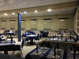 просторный зал ресторана в синих цветах с рядами сервированных столиков у стен, окон с закрытыми жалюзи и по центру