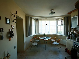 картины, колокольчики, подкова на крючке, веночек на стене светлой кухни с обеденным столом у окна в трехкомнатной квартире геолога
