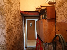 современная деревянная дверь санузла с указателем в коридоре с новым линолеумом из дверного проема прихожей со старыми обоями на стенах, пылесосом у трюмо,шкафами советского времени