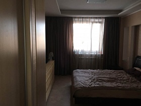 общий вид бежевой спальной комнаты с коричневыми шторами на окне, большой кроватью со светлым покрывалом напротив бежевого комода из открытой двери трехкомнатной квартиры