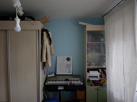 бежевый шкаф для одежды, синтезатор и книги в шкафу за стеклянными дверцами у голубой стены детской комнаты стильной трехкомнатной квартиры