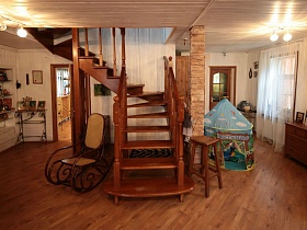 плетенное кресло-качалка, барный стул, детский домик у витой деревянной лестницы рядом с колоной, декорированной плиткой под дикий камень в светлой уютной гостиной семейной классической дачи