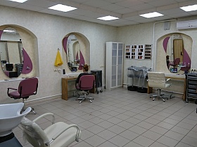 оригинальный дизайн просторного светлого салона парикмахерской 2 с сиреневыми креслами для посетителей у овальных зеркал в нише рабочего места мастера
