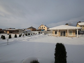 соседние дома за пределами просторного участка загородного коттеджа с одноэтажным светлым домиком с терассой, качелями, светильниками у расчищенной от снега дорожки