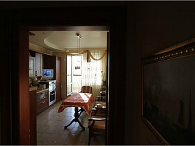 картина на стене у входной двери на кухню с обеденным столом и скатертью напротив окна и двери на балкон  с белой гардиной простой просторной двухкомнатной квартиры