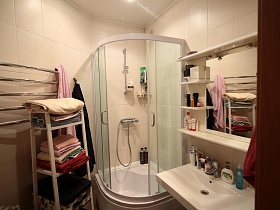 полотенцесушитель на стене, белая этажерка со стопками полотенец ,душевой бокс, зеркало с полками над раковиной в кремовой ванной квартиры художника в сталинке