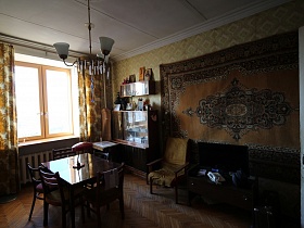 белый телефон и телевизор на тумбочке,кресло с подлокотниками у стены с большим советским ковром на стене,иконы на книжной полке, сервант с посудой у окна гостиной с цветными шторами с желтым рисунком