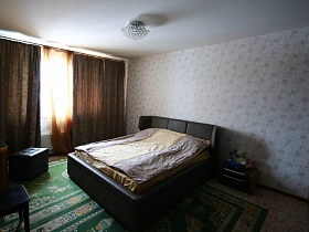 двухцветный гримерный столик у большого окна, прикроватная тумбочка у мягкой кровати с подушками в светлой спальне с зелеными коврами на полу простой квартиры в Химках