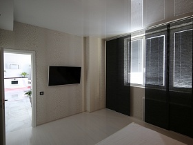 белая кухня через открытую дверь черно белой спальни с плоским телевизором на стене и черными вертикальными жалюзи на окне