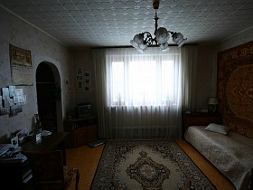 серебристый телевизор на тумбочке и настольная лампа на столе в углу бабушкиной комнаты у большого окна с белой гардиной с ковром на полу и на стене времен 1905-2005 гг яркой трехкомнатной квартиры