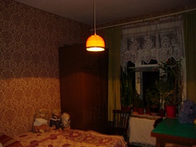 куклы на цветном покрывале кровати, шкаф для одежды в углу у  окна с желтыми шторами и белой гардиной, многочисленные комнатные цветы на подоконнике большого окна спальной комнаты с бежевыми обоями на стенах