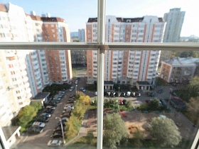 вид из окна квартиры на двор жилых многоэтажек с зеленым участком, детской площадкой и припаркованными машинами вдоль дорог