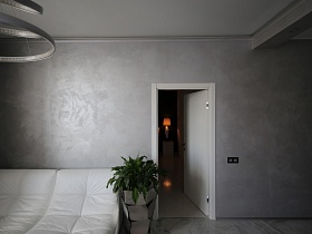 открытая дверь в гостиную с серыми стенами, с серыми кольцами люстрой, серым из пластин вазоне с комнатным цветком у белого мягкого дивана в стильной современной квартиры