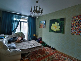 белый мягкий угловой разложенный диван на полу с красными коврами салатовой спальной комнаты с синими шторами на окне и яркими картинами на стенах лофт квартиры