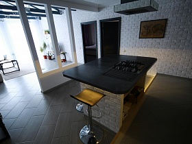 барные стулья на металлической круглой ножке вокруг обеденного стола-шкафа со встроенной газовой плитой в черной столешнице в зоне кухни загородного домика для съемок кино