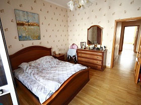 большая  деревянная кровать со светлым постельным , шкаф  для одежды и комод в светлой спальной комнате с открытой дверью простой большой квартиры в Марьино