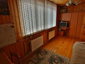 шкаф для одежды, небольшой телевизор на встроенном столе под навесным шкафчиком у окна, ковровые дорожки у кровати в детской комнате деревянной загородной дачи