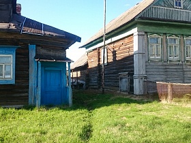 голубая входная дверь крыльца  перилами деревянного дома-сруб с голубыми наличниками на окнах по соседству с открытым балконом с перилами на втором этаже дома на высоком цоколе