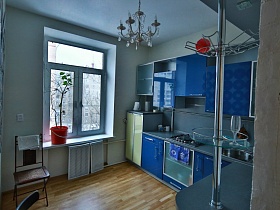 синяя мебельная стенка с барной фурнитурой над голубой столешницей шкафа в кухне с люстрой, стилизованной под свечи на белом потолке современной лофт квартиры