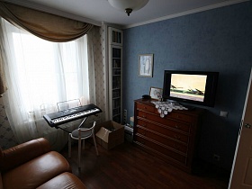 синтезатор и стул у окна гостиной с белой гардиной и бежевыми короткими шторами, встроенный шкаф с книгами в семейной трешке