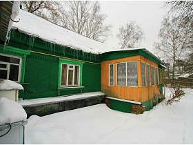 сосульки свисают с крыши зеленой дачи с пристроенной деревянной верандой в зимнее время года