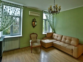 настенные часы над бежевым креслом и угловой бежевый мягкий диван у окна с прозрачными зелеными гардинами в салатовой гостиной трехкомнатной квартиры №16