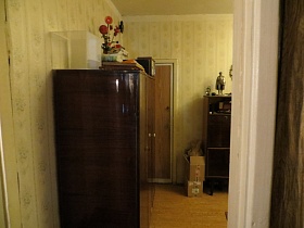 стеклянный пустой аквариум, часы, ваза с цветами на поверхности полированного шкафа у стены с полосатыми светлыми обоями гостиной комнаты из открытой двери двухкомнатной квартиры молодой советской семьи
