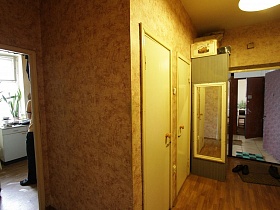 ящики и коробки на верху и прямоугольное зеркало на боковой поверхности серого шкафа для одежды, старые двери санкомнат в прихожей с бежевыми обоями бедной квартиры жилого дома